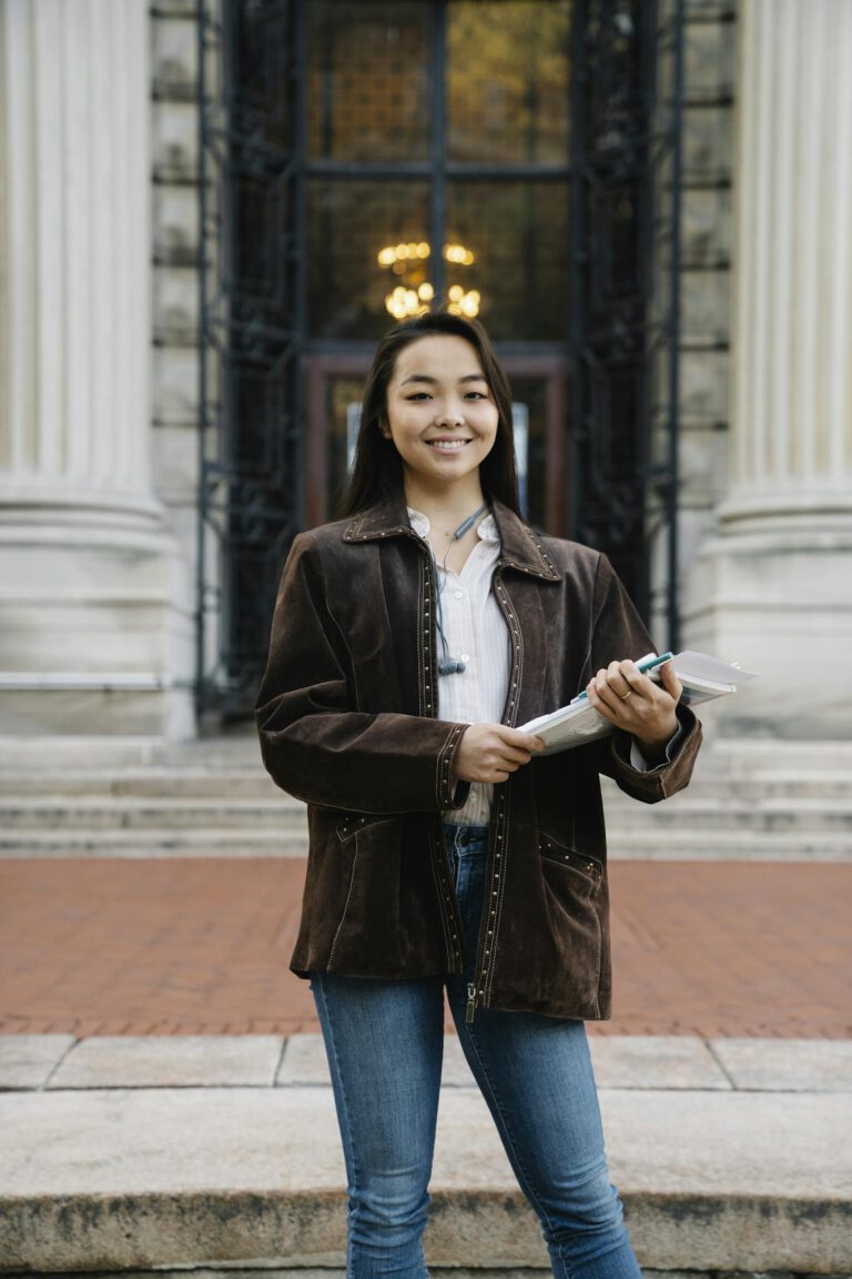 Smiling Asian woman holding documents standing in front of an ornate building.