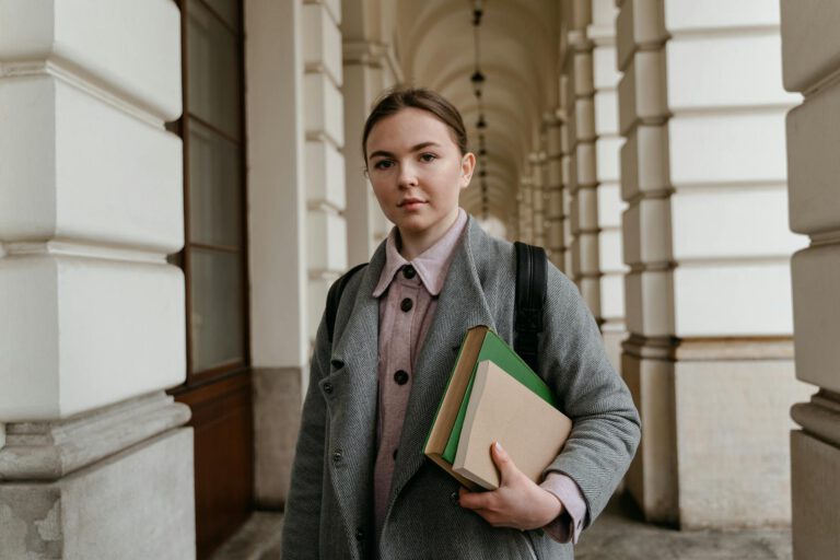 Young woman carrying books in an architectural hallway, dressed for fall weather.